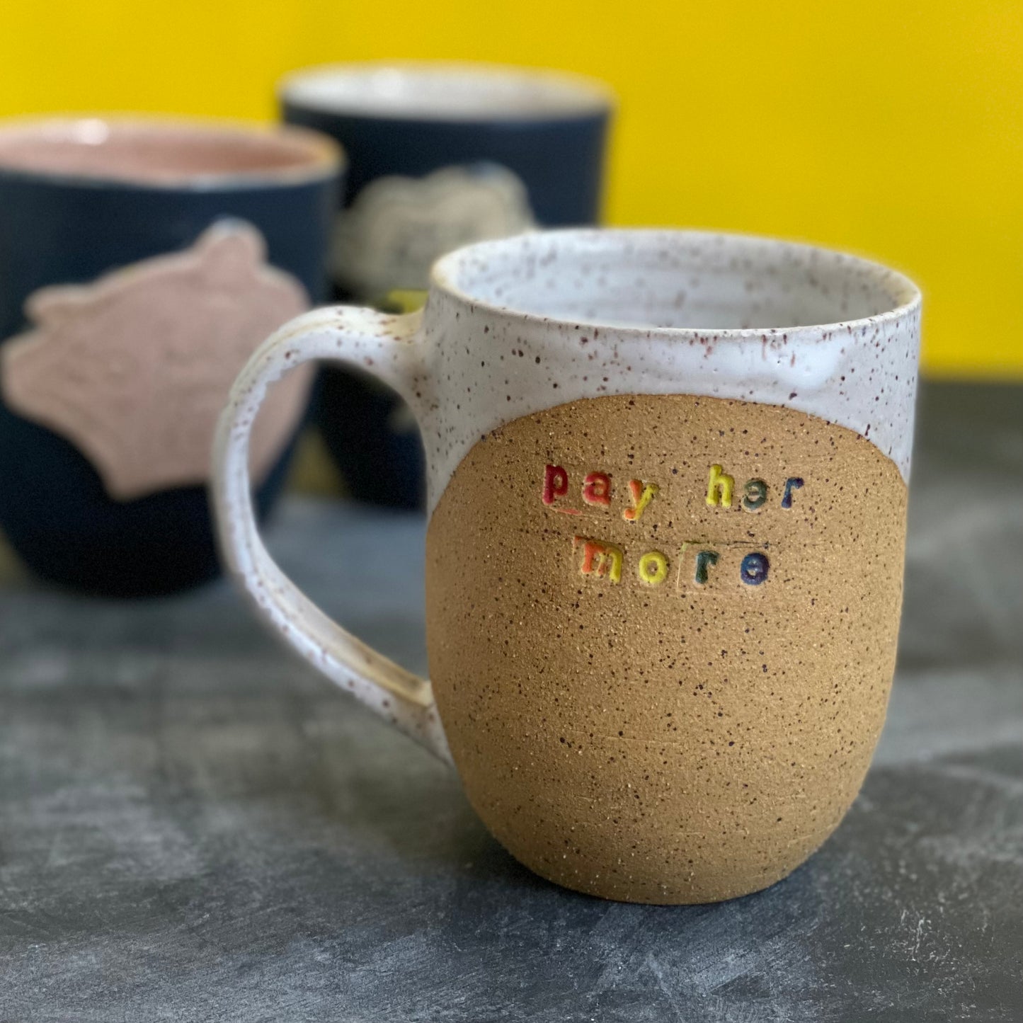 pay her more mug with white glaze and rainbow text