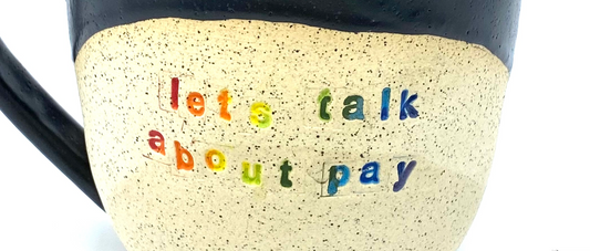 closeup of rainbow text "lets talk about pay" on a cream mug with black speckles