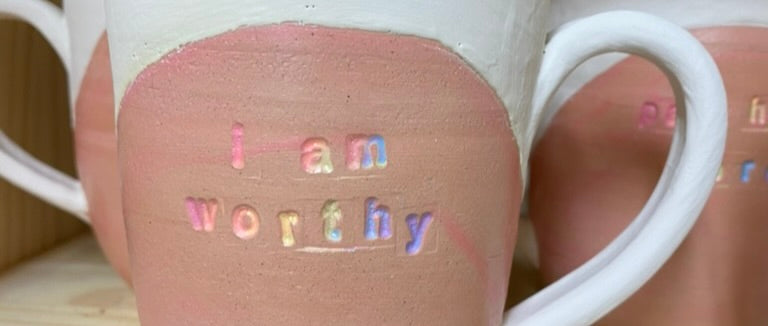 a mug in process, the rainbow words "i am worthy" covered in pink wax below a white glazed rim and handle