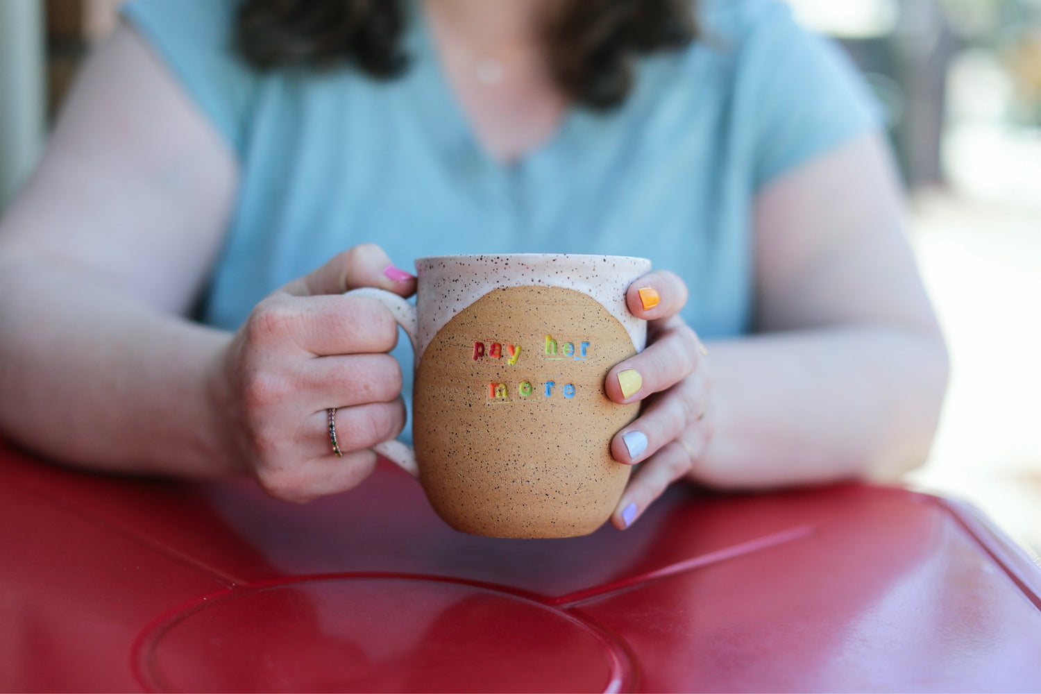 "pay her more" mug held in hands wearing rainbow ring and rainbow painted nails. photo by andrea scher