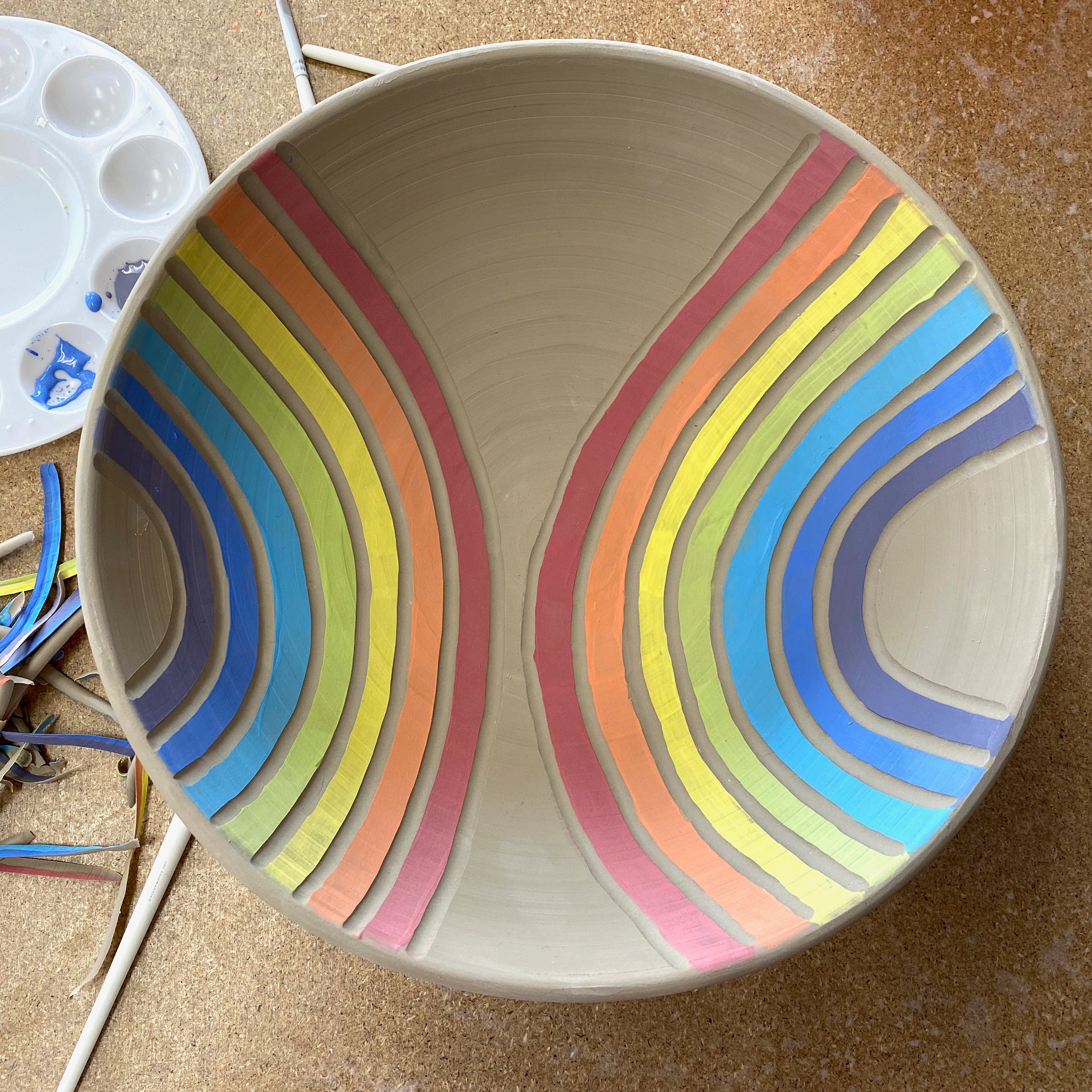 A greenware porcelain bowl seen from above, decorated with painted and carved rainbows.
